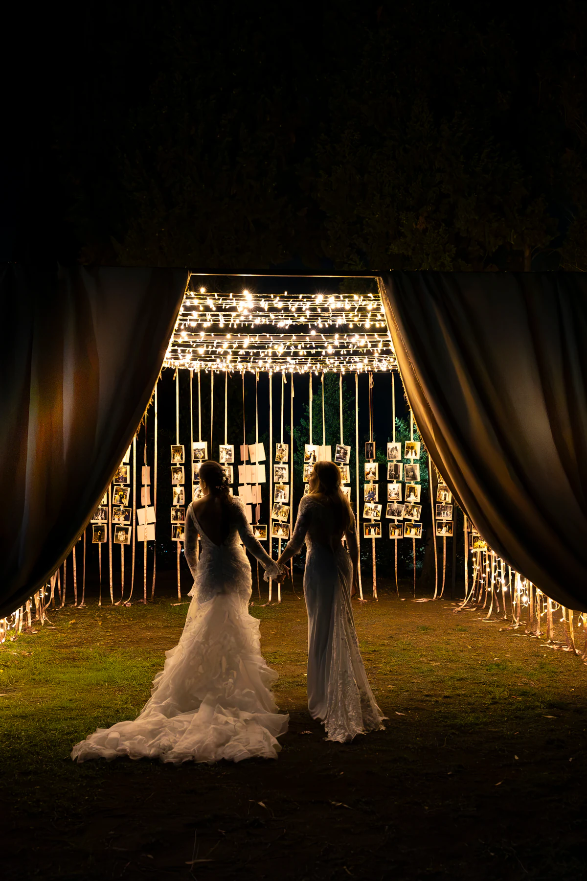 Two brides walking hand in hand into the Light Gallery, between open curtains and strings of light