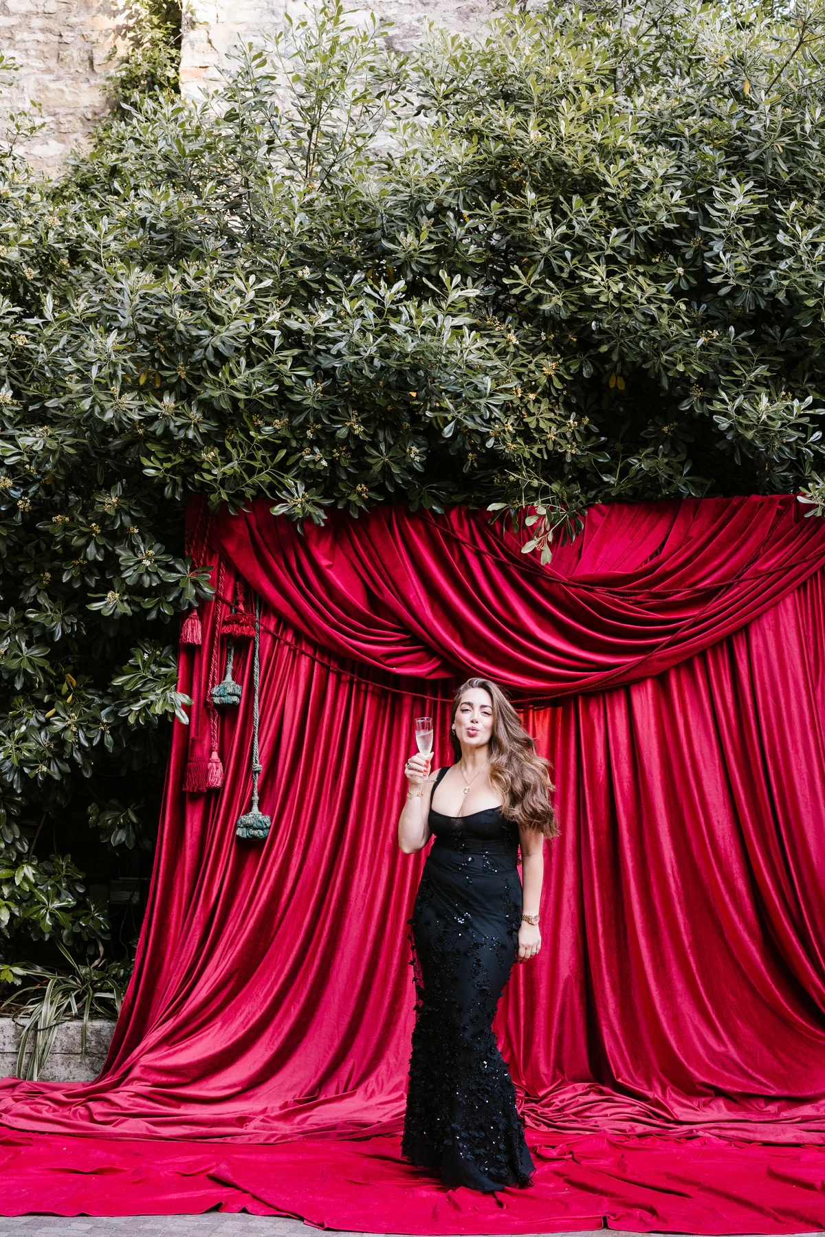 A guest in a black gown with a glass of champagne, posing in front of the red velvet curtain