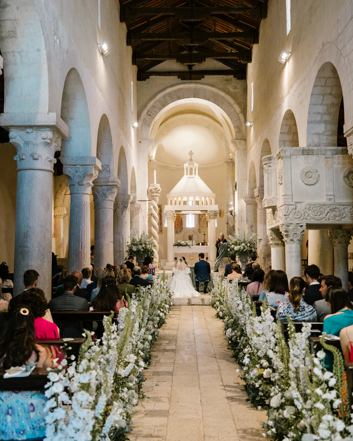 A wedding ceremony in a stone church, the aisle lined with white flowers