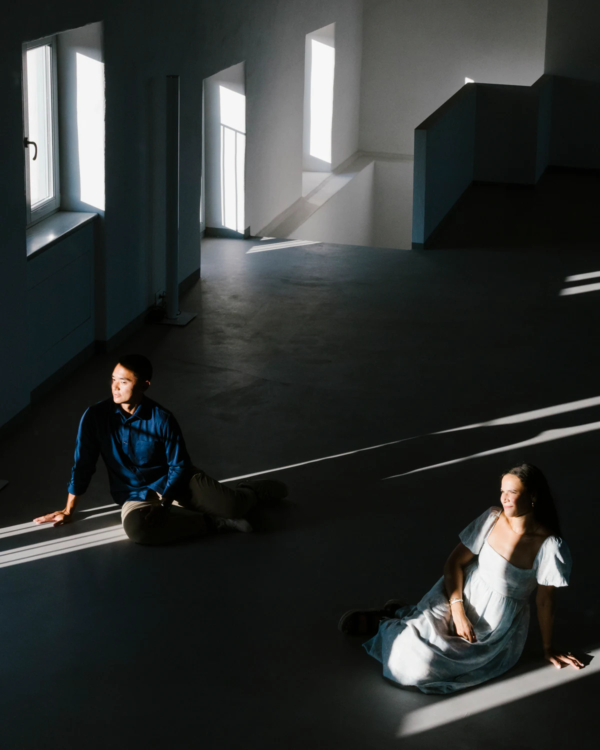 A man and a woman sitting on the floor of a dark room, in shafts of light