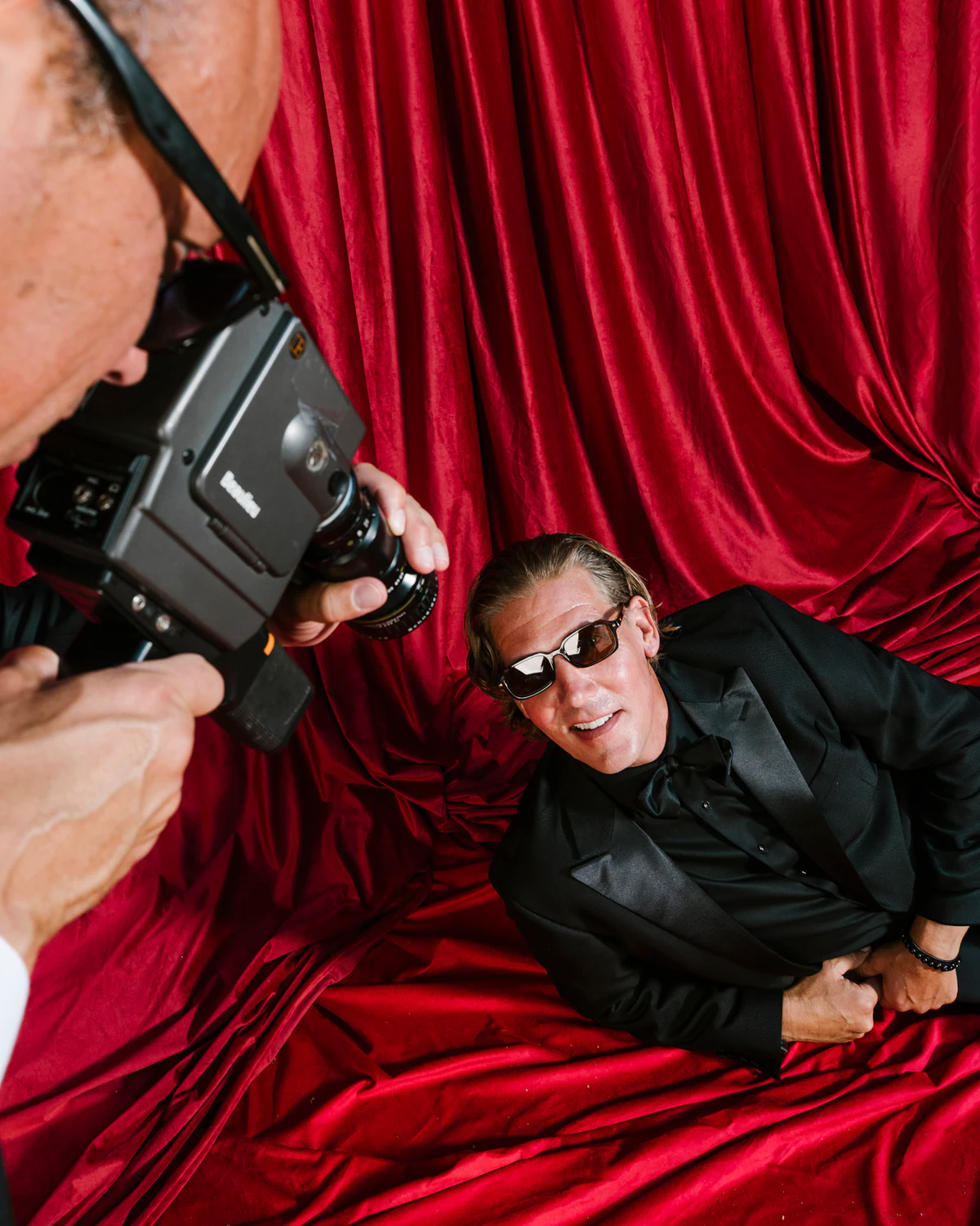 A man filming with a cine camera beside a man in sunglasses lying on red velvet