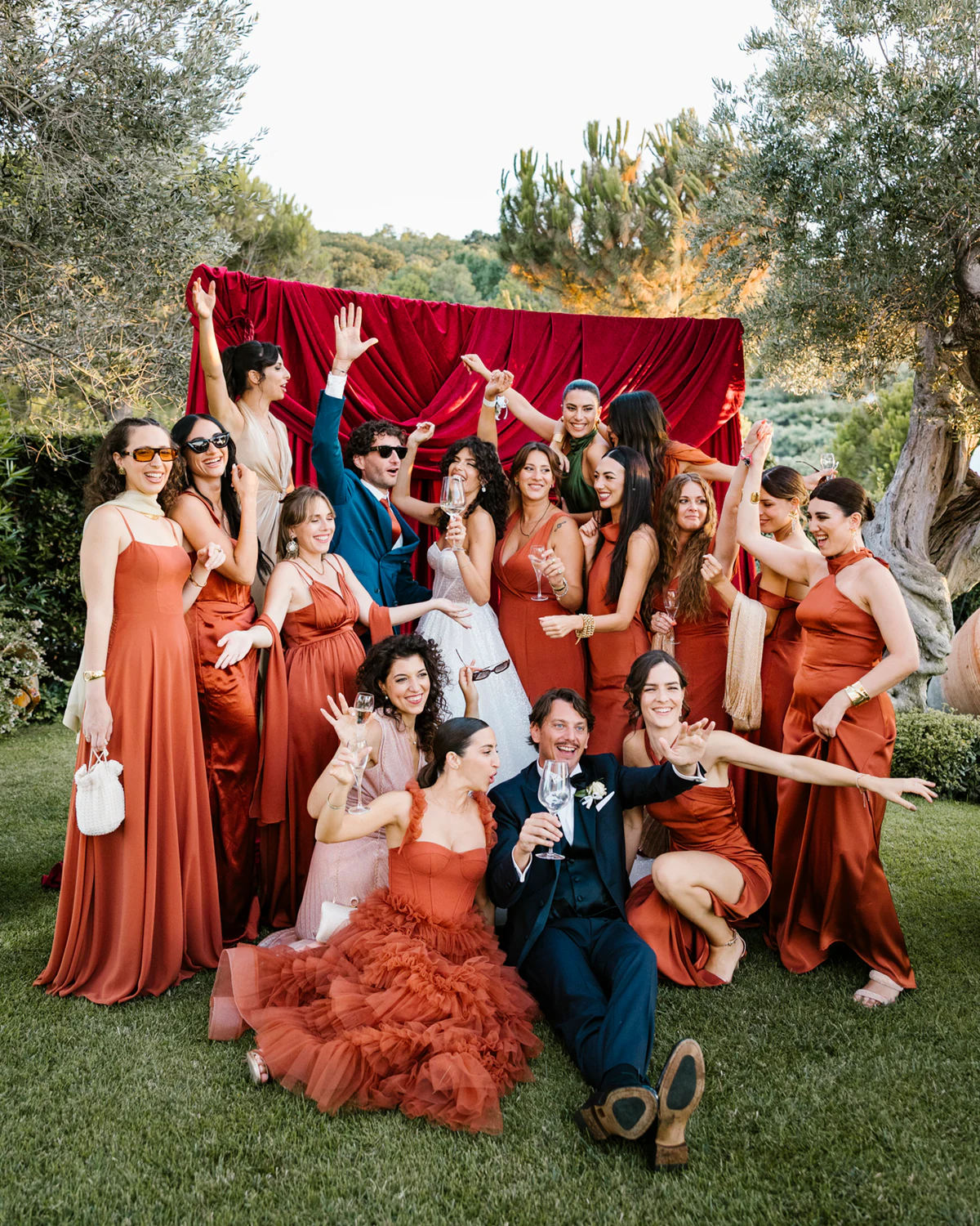A wedding party in terracotta dresses celebrating in front of a red backdrop