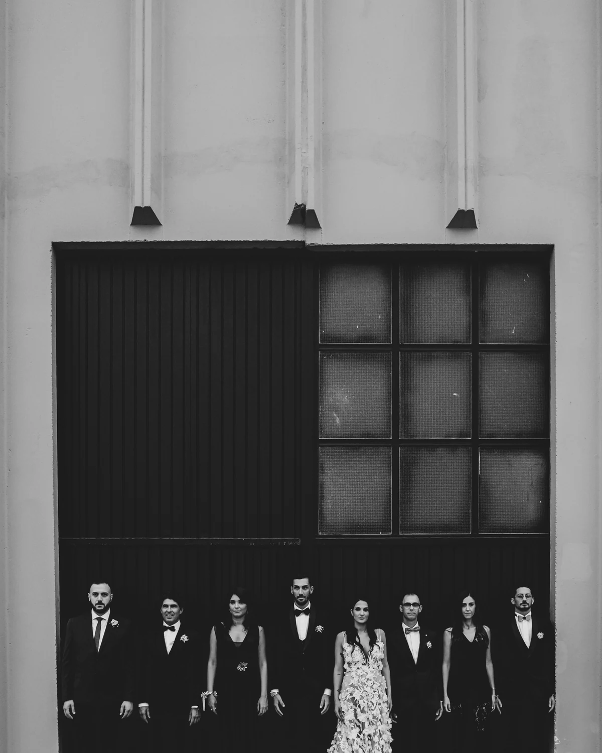 A wedding party standing in a row beneath a tall dark doorway, in black and white