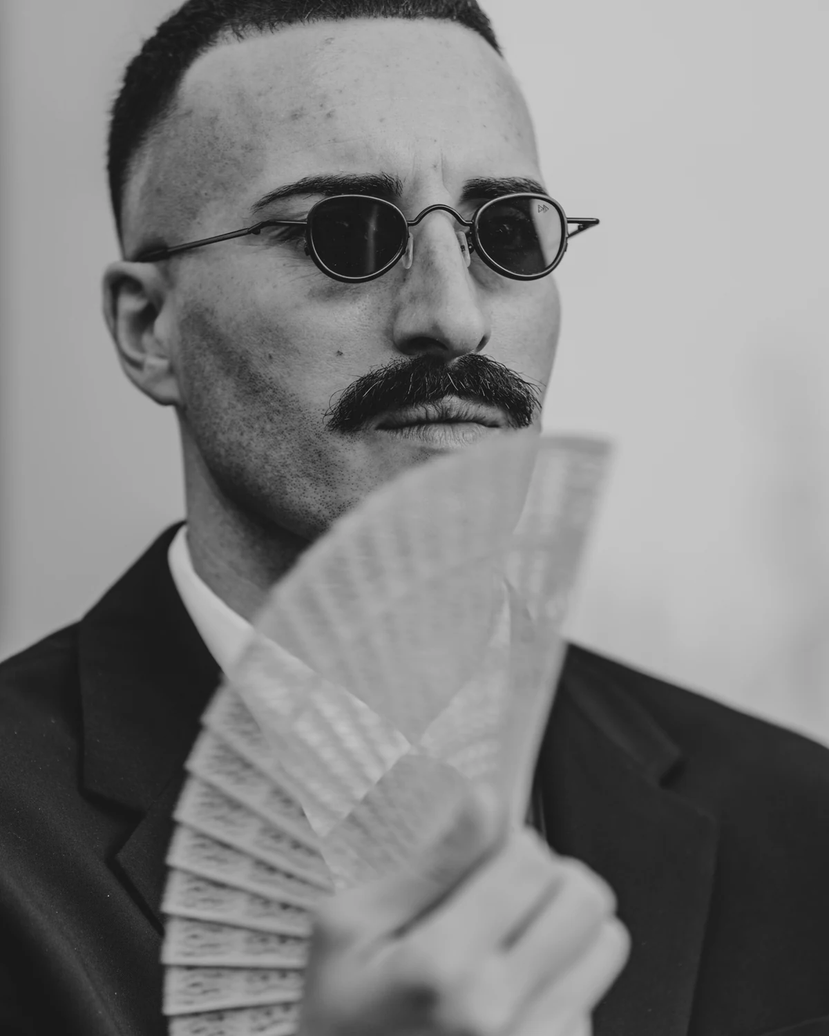 A man with round sunglasses and a moustache holding a fan, in black and white
