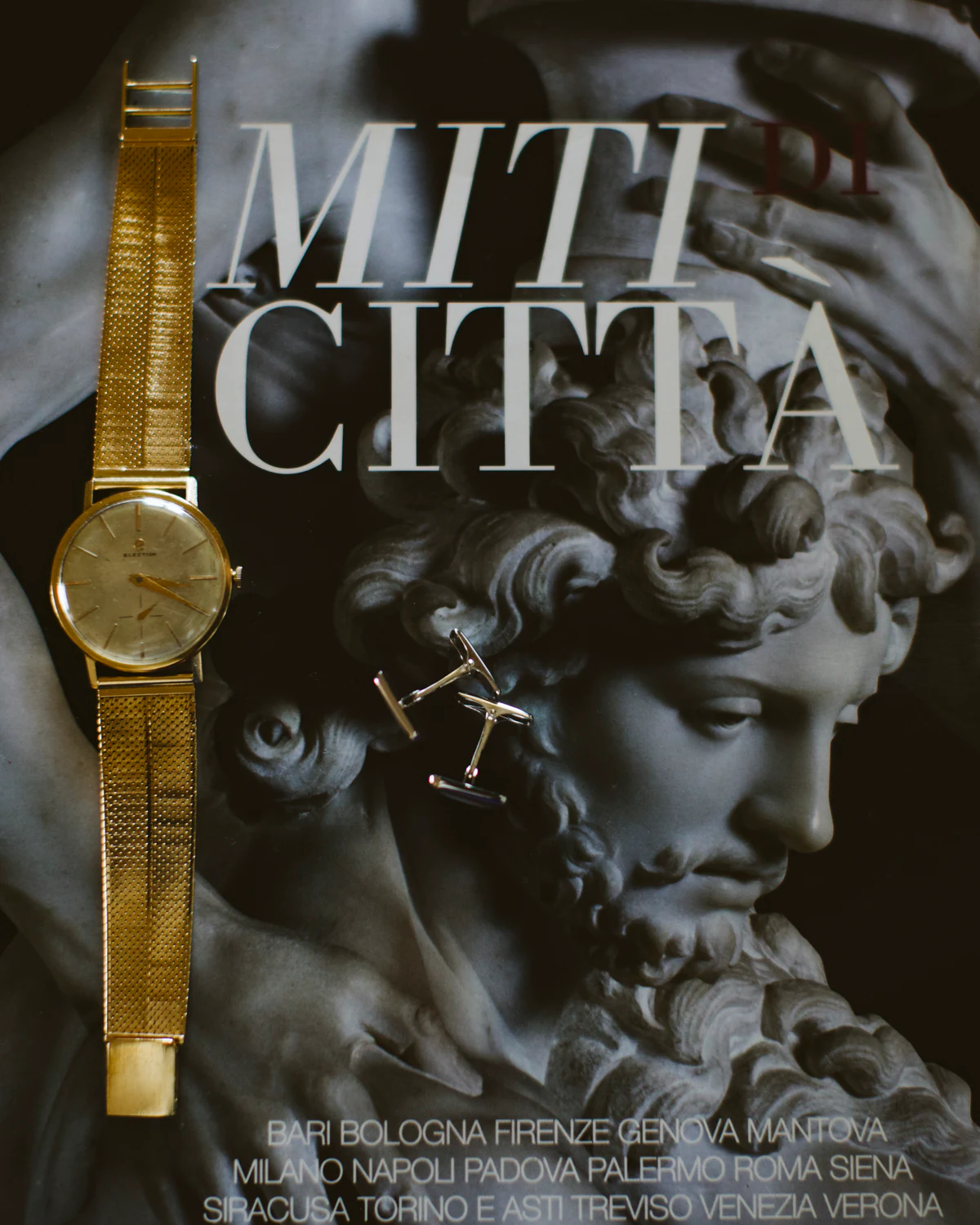 A gold watch on a magazine page titled Miti Città, beside a classical sculpture