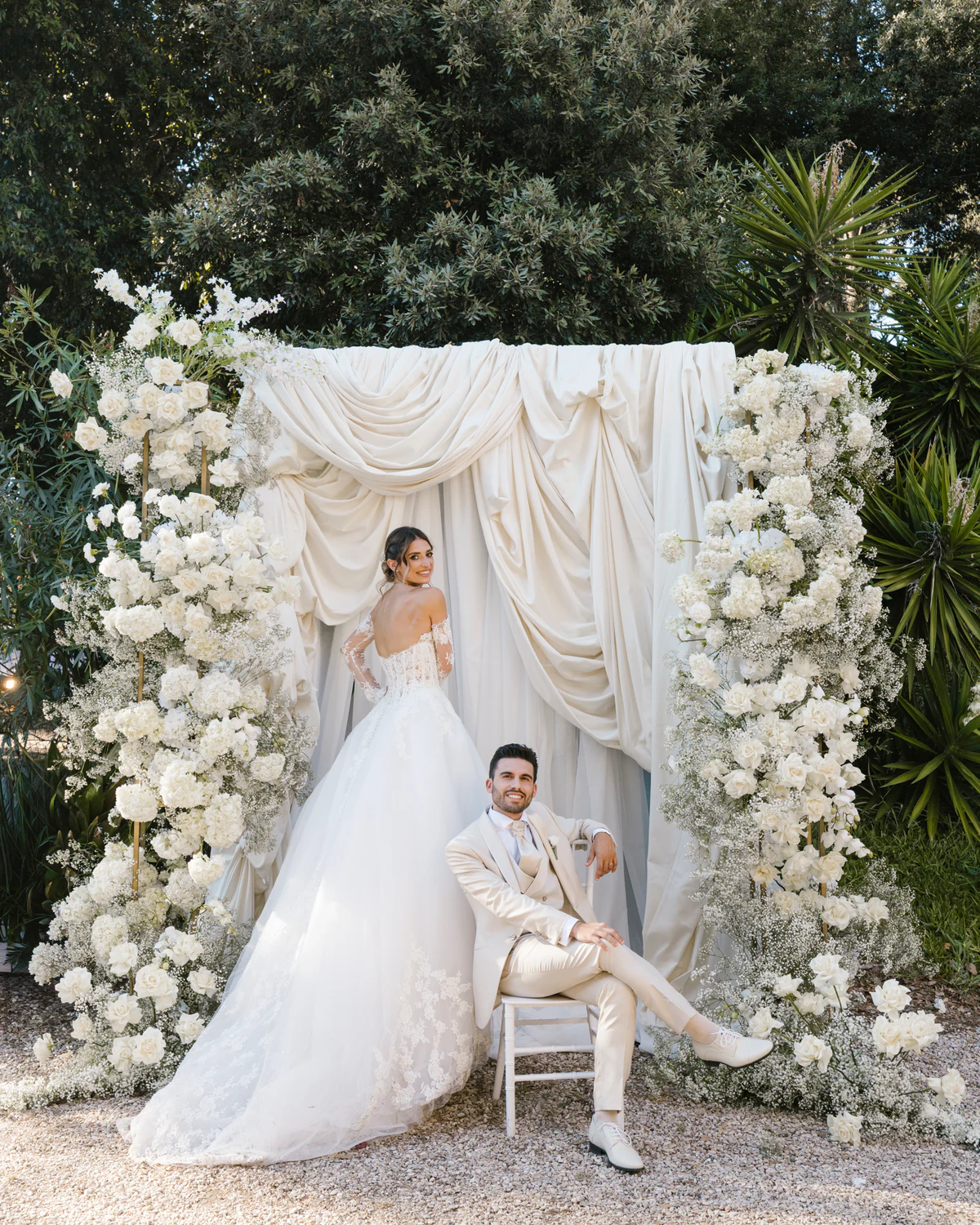 A bride and a groom in a cream suit beneath an arch of white flowers