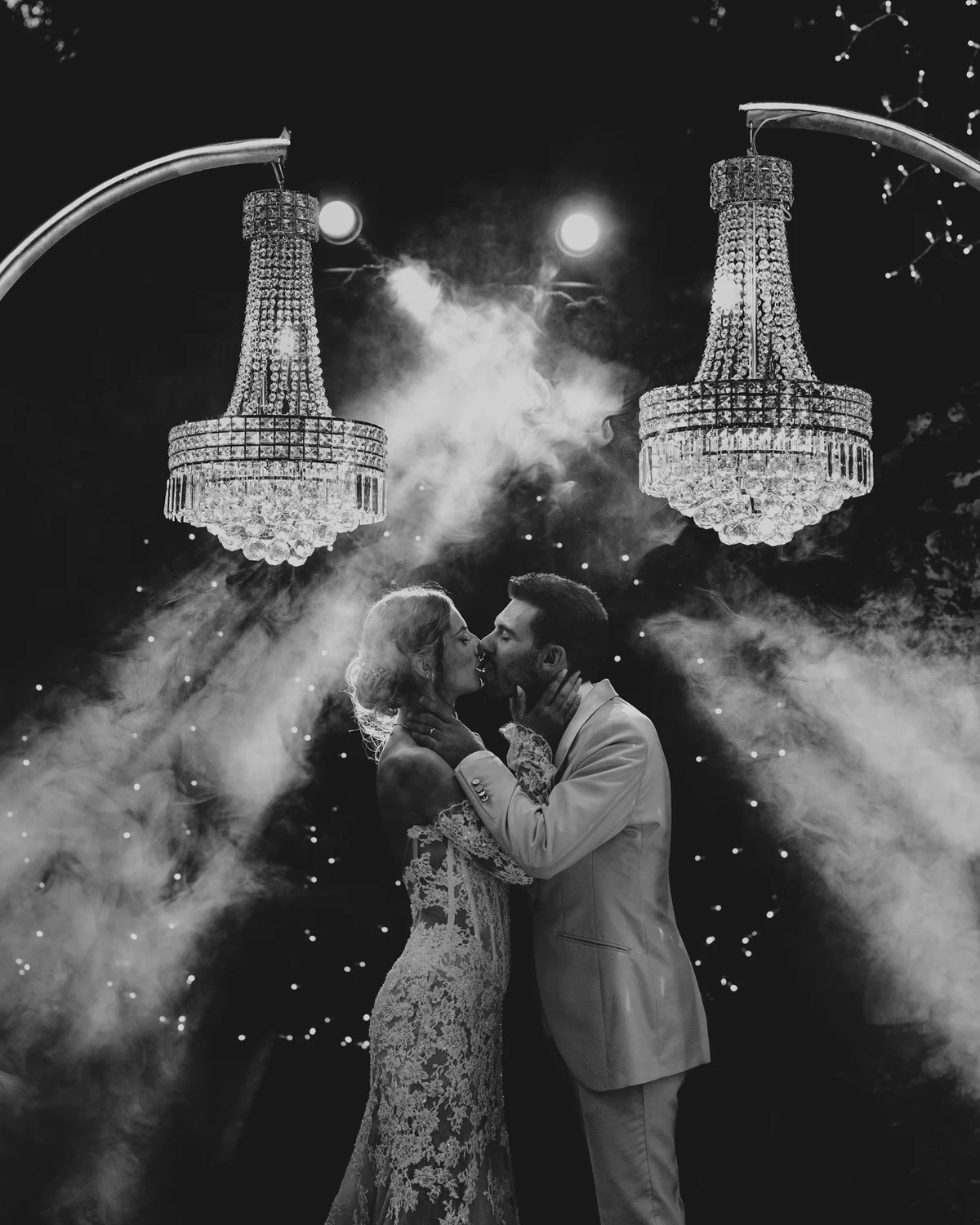 A couple kissing beneath two crystal chandeliers in a haze of smoke, in black and white