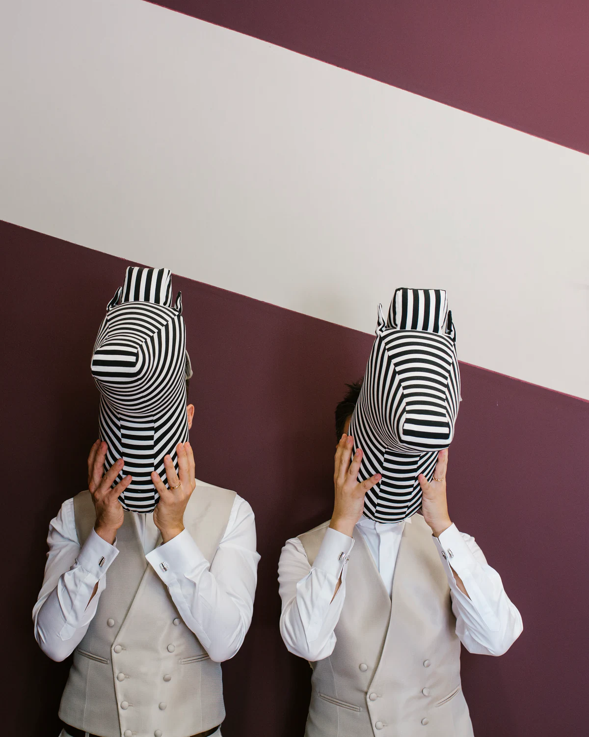 Two people holding striped masks in front of their faces against a two-tone wall