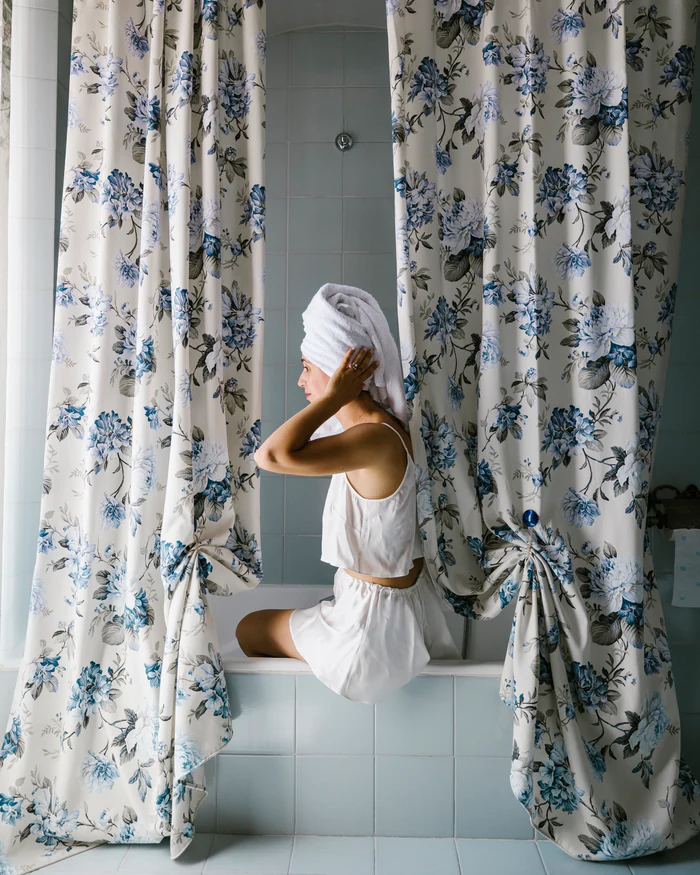 A woman with a towel wrapped around her head, sitting on the edge of a bathtub between floral curtains