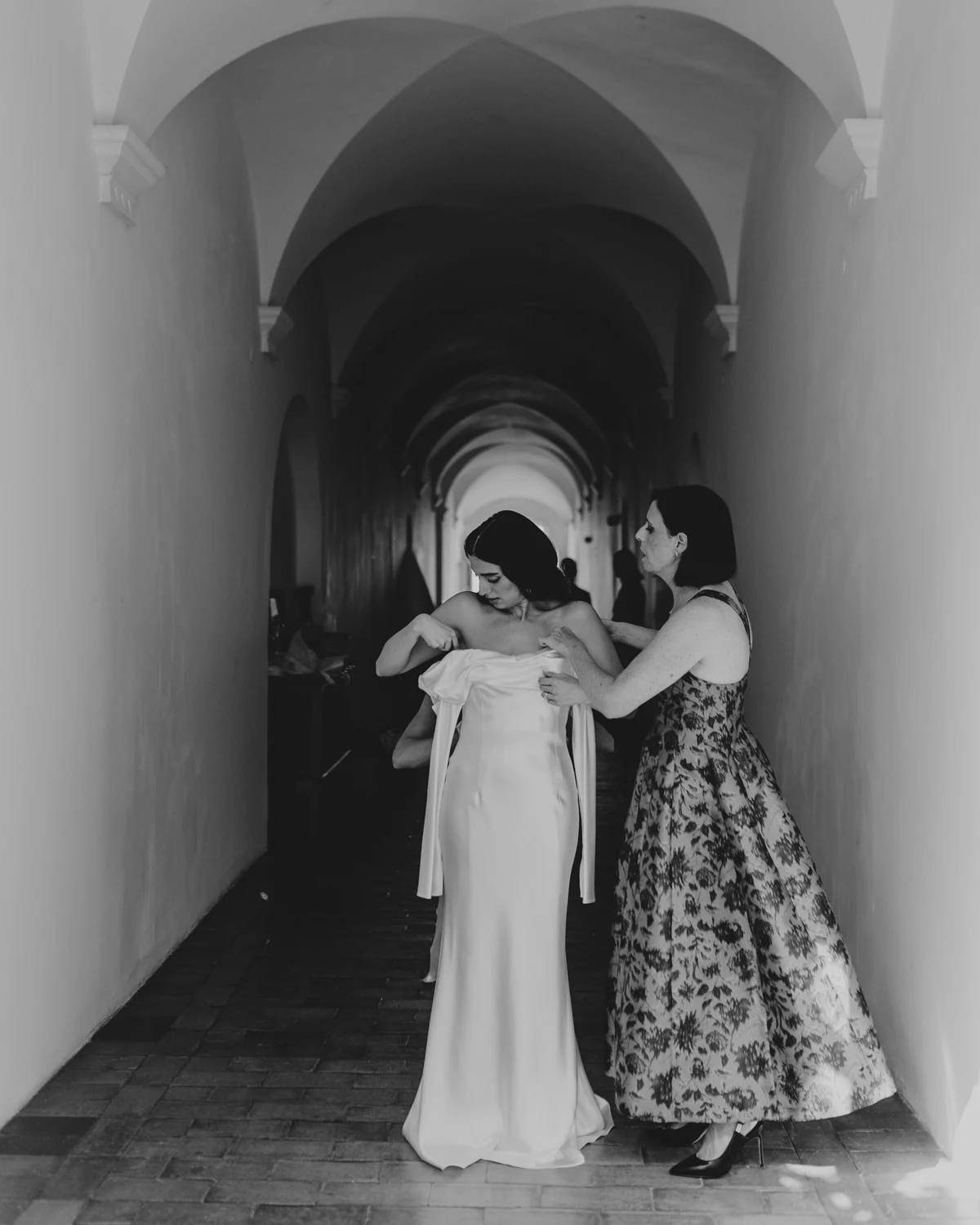 The bride embraced by a woman in a floral dress, in a vaulted corridor