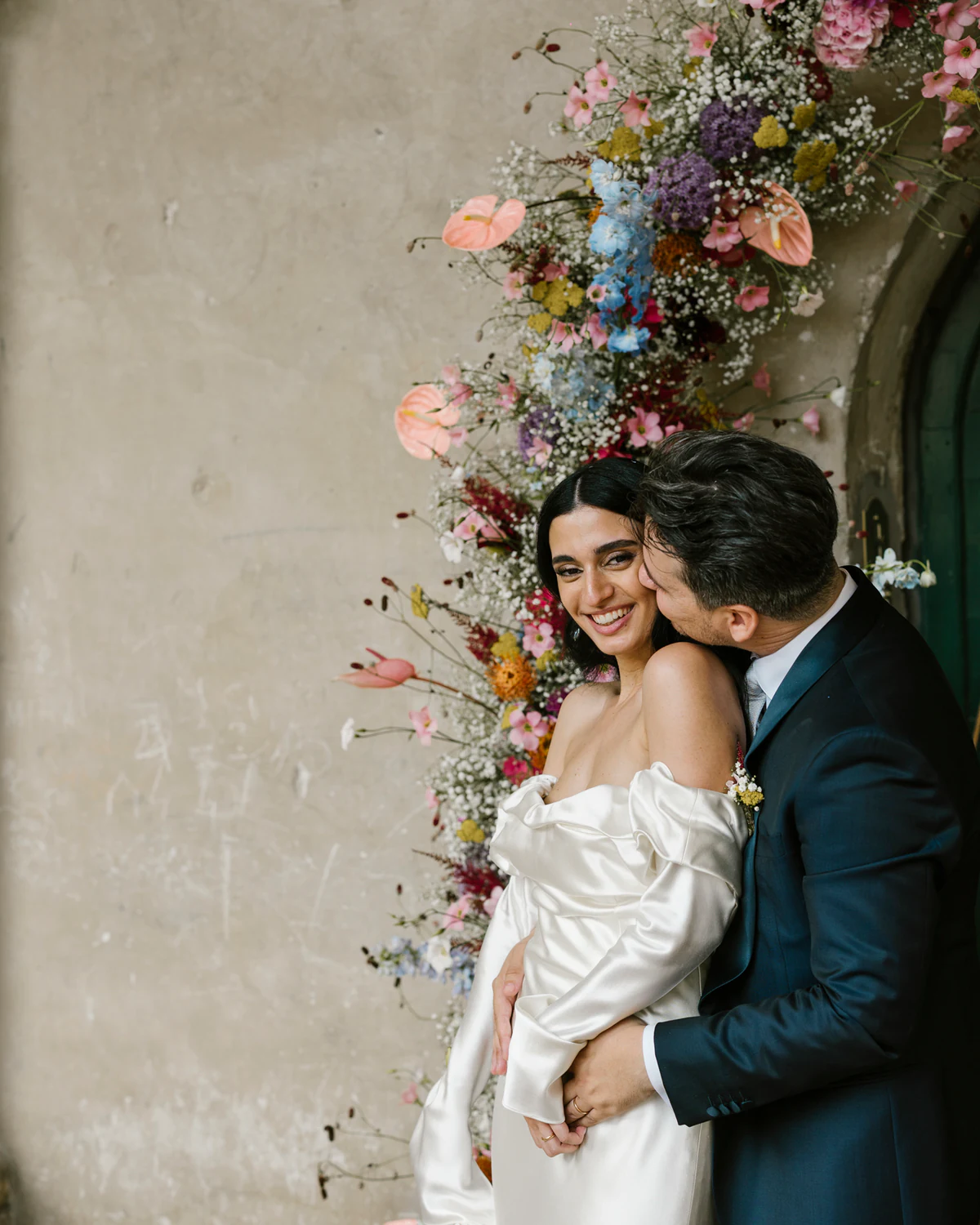 A bride and groom embracing beside an arch of flowers, the bride smiling at the camera