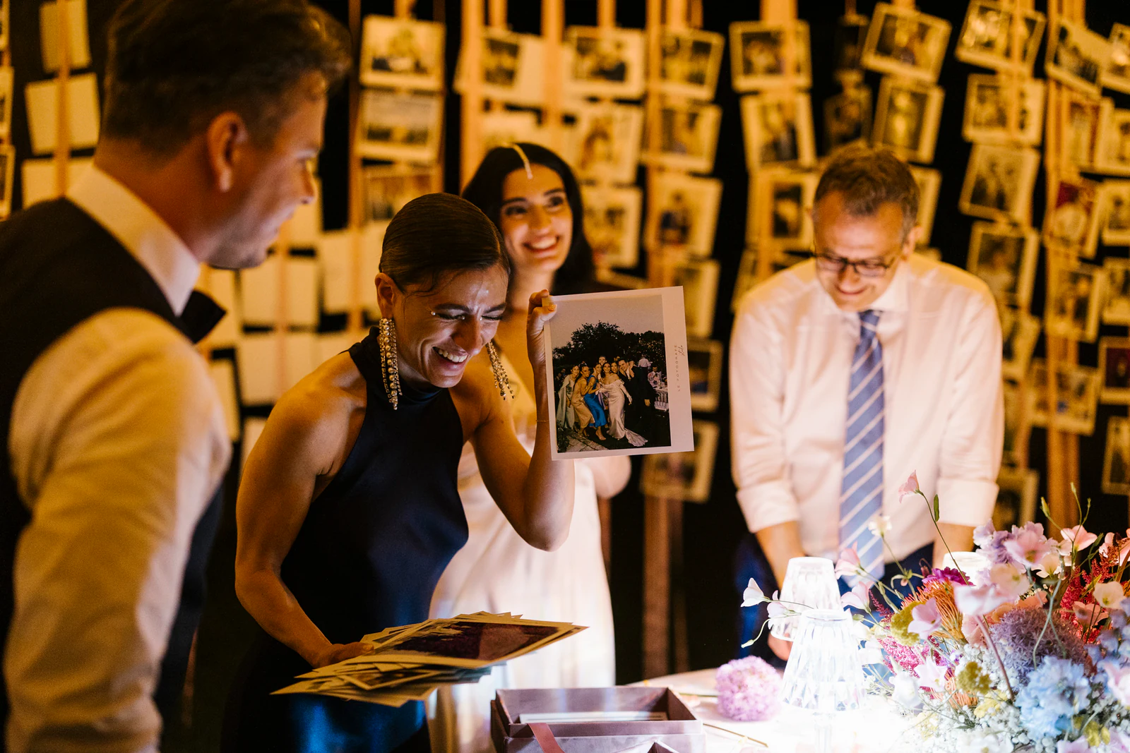 The bride and her guests laughing over photographs, in front of a wall of pictures