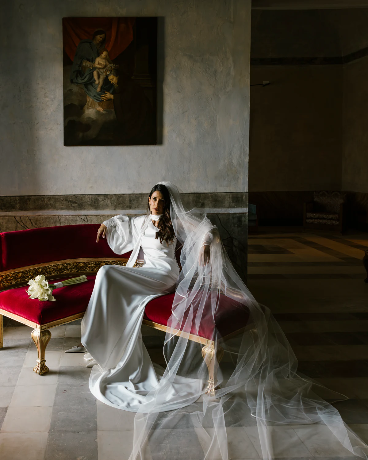 The bride seated on a red sofa, her long veil spread on the floor