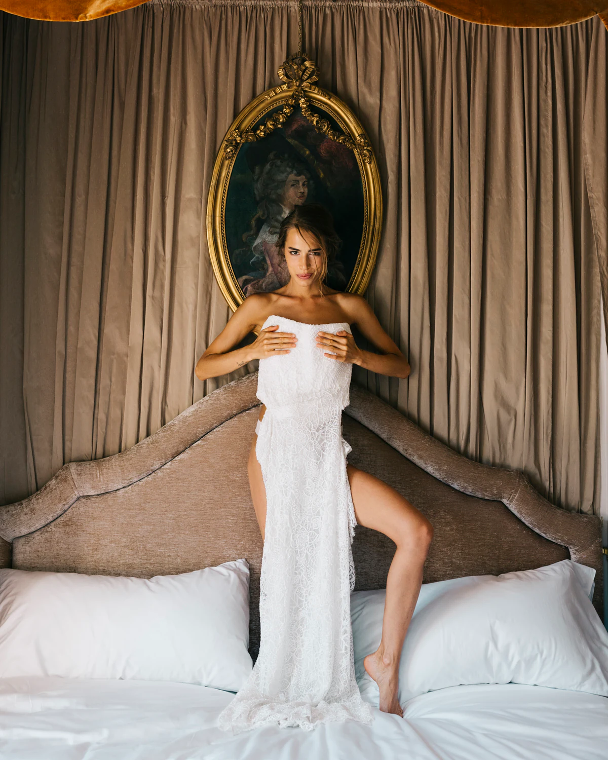 The bride on the bed holding her dress, under a gilded mirror