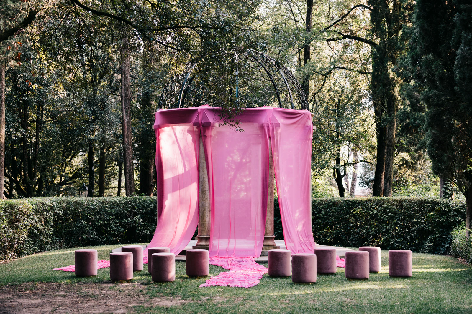 A gazebo draped in pink silk among the trees, with pink stools