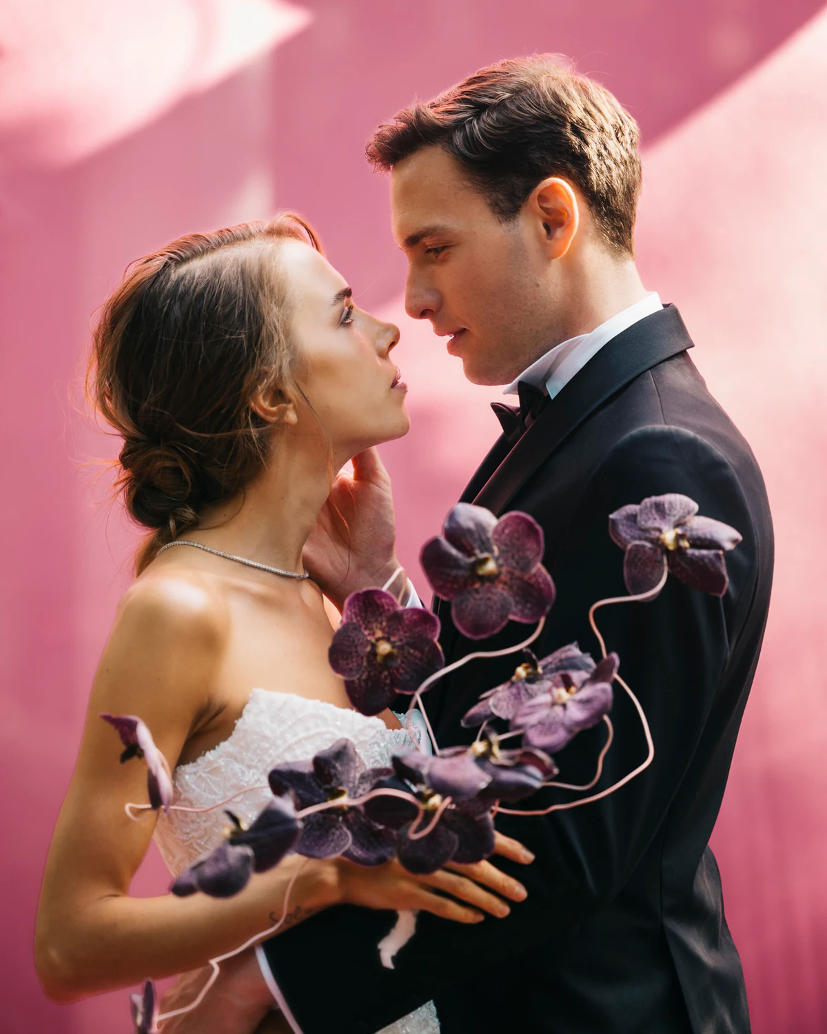 The couple face to face against pink silk, with orchids