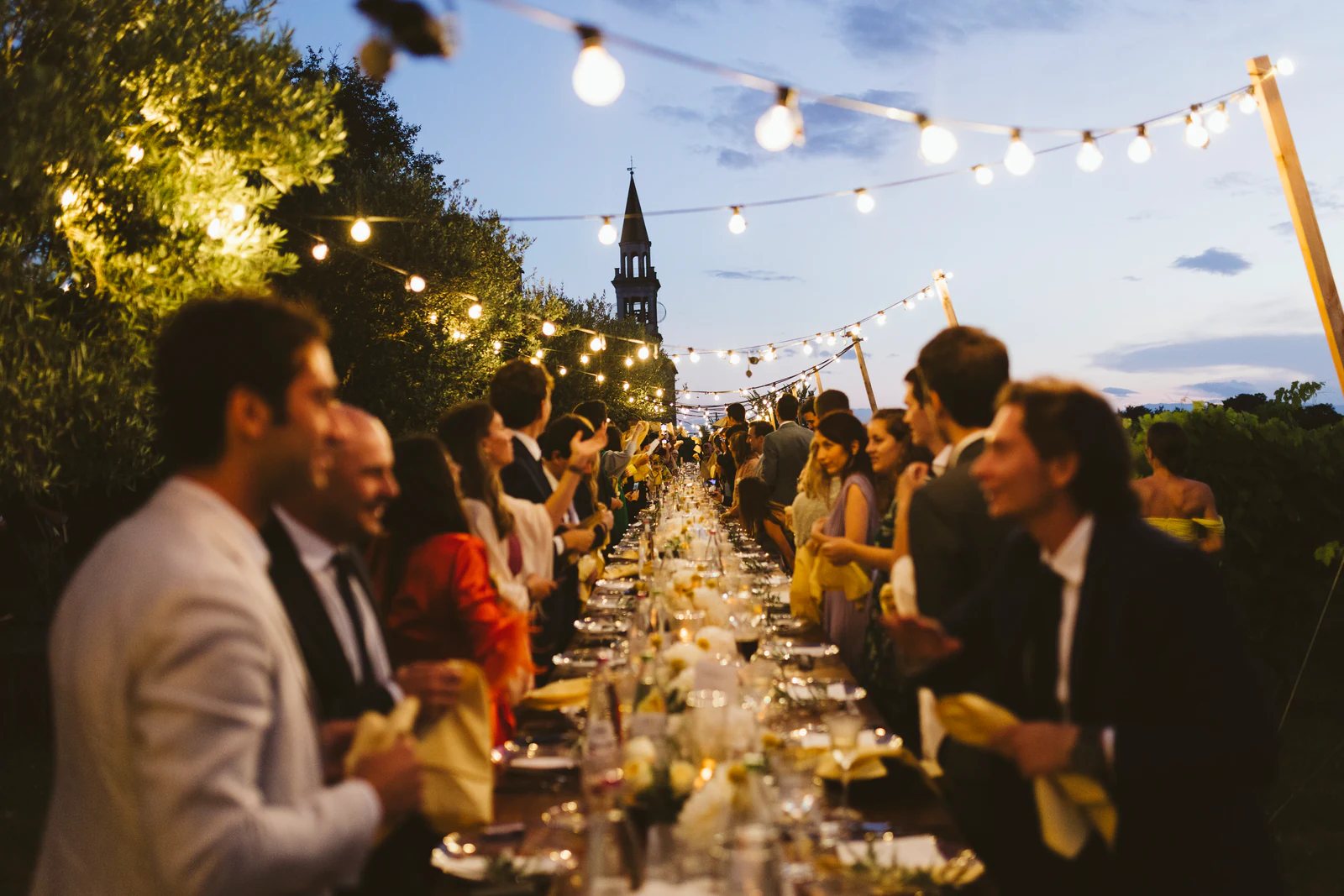 Guests at the long table under string lights, the bell tower behind